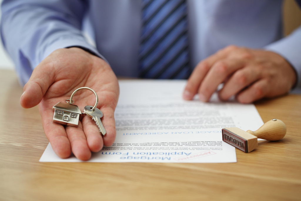 a real estate agent holding a house key with a house keychain on one hand and touching a paper with the other hand on top of a table beside a stamp
